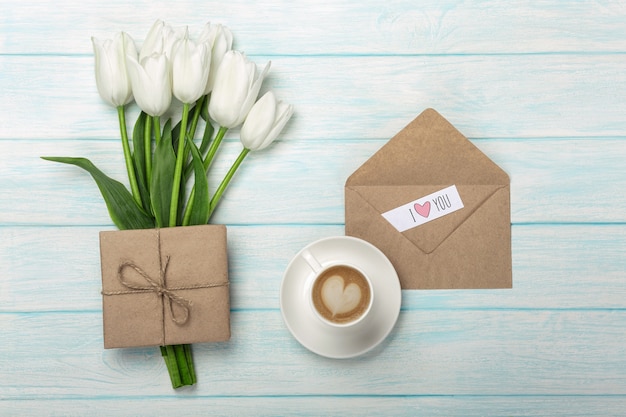 Een boeket van witte tulpen, kopje koffie met een liefdesbrief en envelop op blauwe houten planken