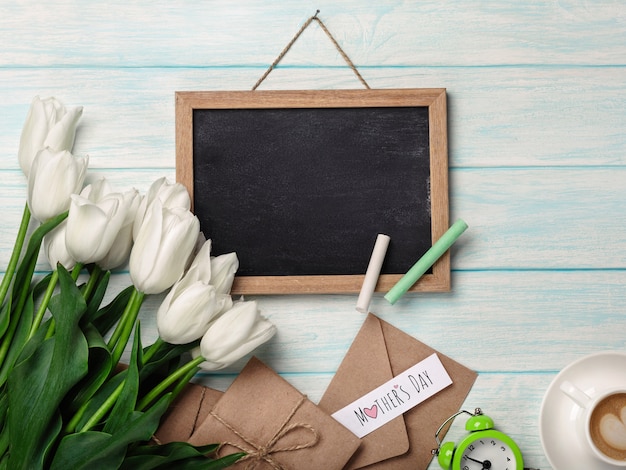 Een boeket van witte tulpen met krijtbord, kopje koffie, liefdesbrief en enveloppen op blauwe houten planken. moederdag