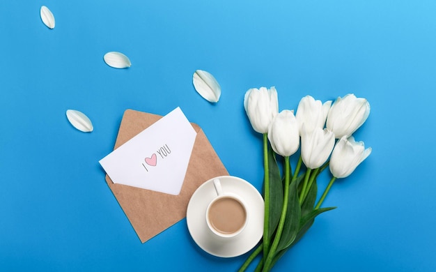 Een boeket witte tulpen en een kopje koffie met een liefdesbriefje op een blauwe achtergrond