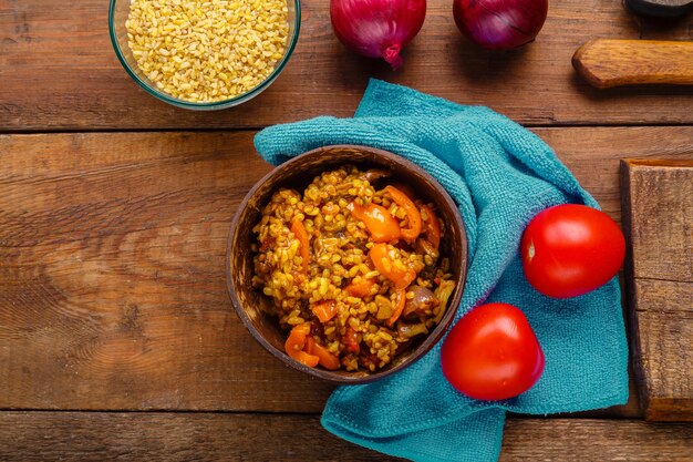 Een bord bulgur met groenten en champignons naast uien en tomaten en een kopje bulgur op een houten tafel