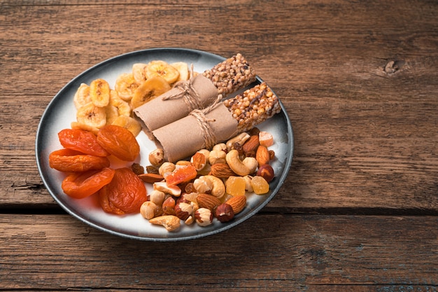 Een bord gezonde zoete snacks. Gedroogde abrikozen, bananenchips, noten en mueslireep op een houten muur. Zijaanzicht, kopieer ruimte.