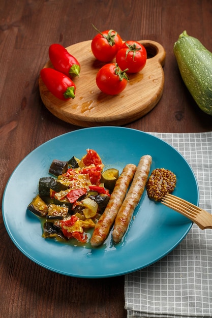 Een bord groenten met gegrilde worstjes op tafel naast tomaten aubergine op planken en een houten vork