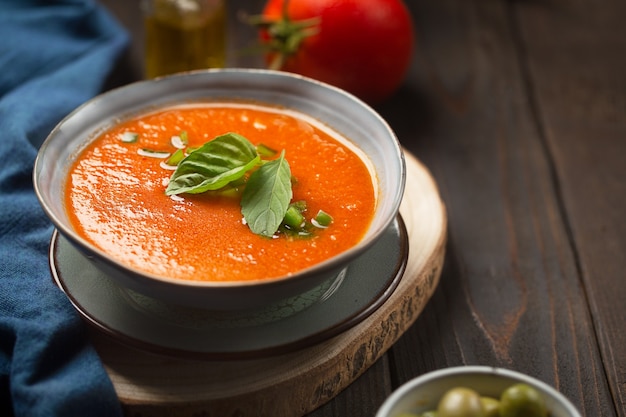 Een bord koude tomatengazpacho, komkommerpepers met tabascosaus op een donkere houten tafel met olijfolie en ingrediënten met een blauw servet. Traditioneel Spaans gerecht