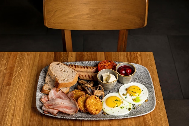 Een bord met groot Engels ontbijt gebakken eieren hash browns spek champignons worstjes gegrilde tomatenbrood boter kersenjam geserveerd op een grijze plaat op een houten tafel van een restaurant
