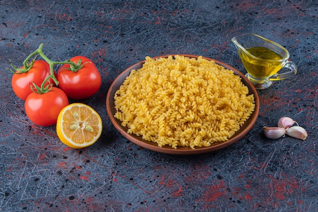 Een bord ongekookte spiraalpasta met olie en verse rode tomaten op een donkere ondergrond.