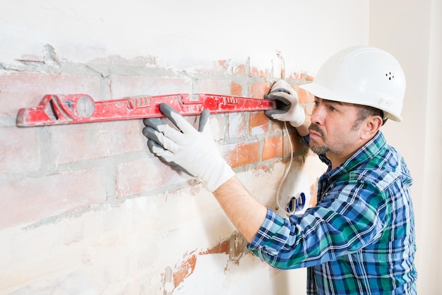 Een bouwer met een veiligheidshelm controleert een platte bakstenen muur met een gebouwniveau tijdens het repareren van een huis