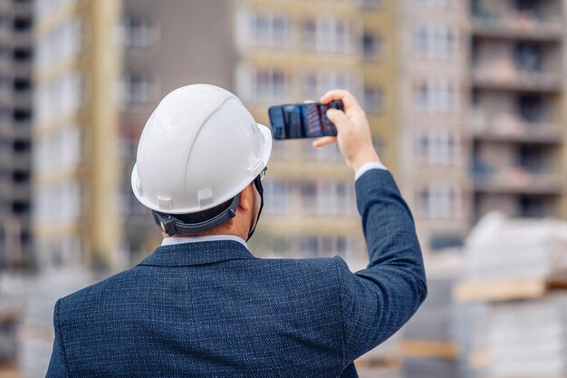Een bouwingenieur met een helm staat tegen de achtergrond van een huis in aanbouw en fotografeert het proces.