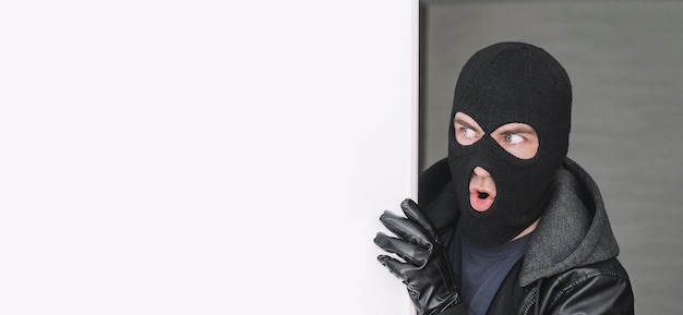 Een boze dief in masker kijkt uit van achter een wit bord Ruimte voor tekst en design kopie ruimte de verraste bandiet kijkt weg