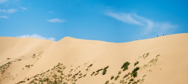 Een breed panorama van een groot duin met een klein silhouet van mensen die erop lopen tegen de blauwe lucht.