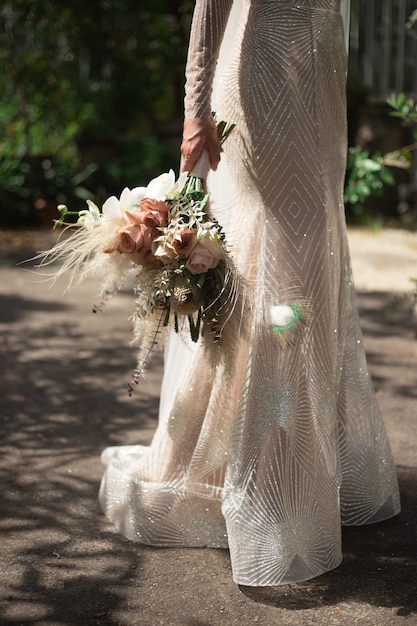 Een bruid in een glanzende trouwjurk staat en heeft een bruidsboeket in boho-stijl in haar hand.