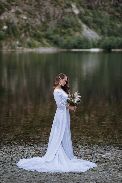 Een bruid in een lange trouwjurk staande op de oever van een meer