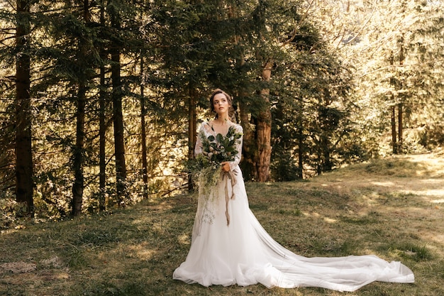 Een bruid in een witte jurk staat in het bos met een mooi bruidsboeket