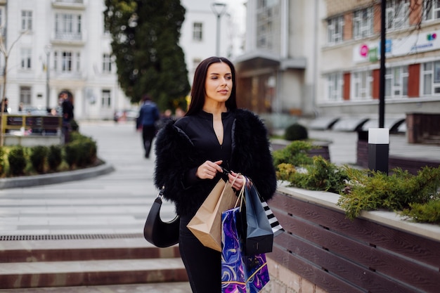 Een bruinharige vrouw, gekleed in zwarte kleding, houdt kleurrijke boodschappentassen vast tijdens een succesvolle shopping spree. Buiten lopend geniet ze van de warmte van een dag