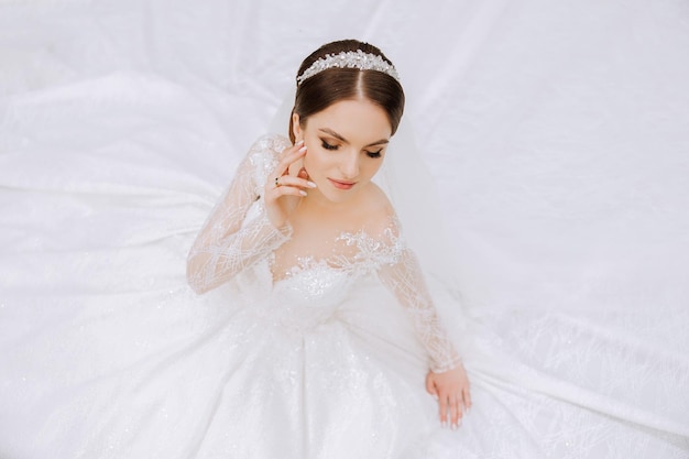 Een brunette bruid met een tiara in een lacy witte jurk met lange mouwen poseert tegen de achtergrond van haar jurk Op een witte achtergrond Klassieke kapsel Avondmake-up Lente bruiloft