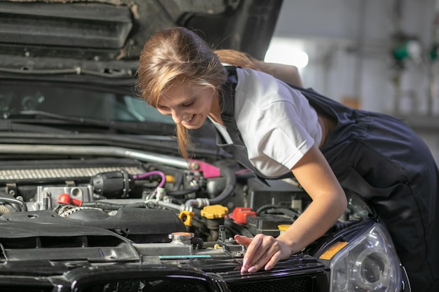 Een brunette in een zwarte jumpsuit en een wit t-shirt bij de open motorkap van een zwarte auto. Jonge vrouw in de garage lacht naar de camera en kijkt neer