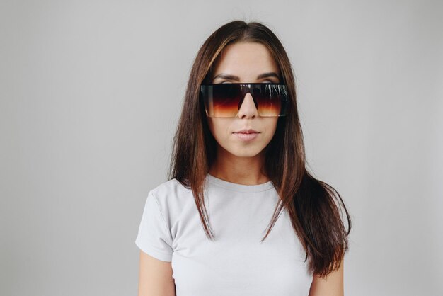 Een brunette meisje in een wit t-shirt op een witte achtergrond staat in een enorme zonnebril