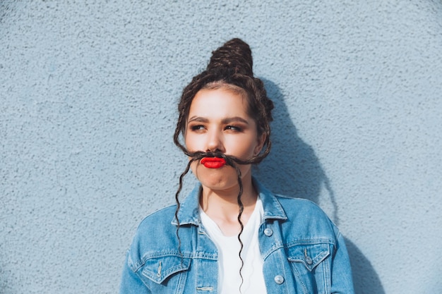 Een brunette meisje met dreadlocks en rode lippenstift in een spijkerpak staat in de buurt van een blauwe muurgeneratie z