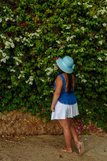 Een brunette vrouw in een witte rok en een zonnehoed staat bij de bloeiende witte bougainvillea