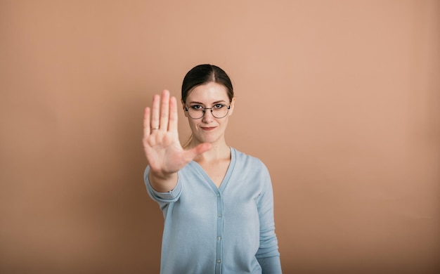 Een brunette vrouw met een bril in een blauwe trui staat en toont een stop op een beige achtergrond met een plek voor tekst