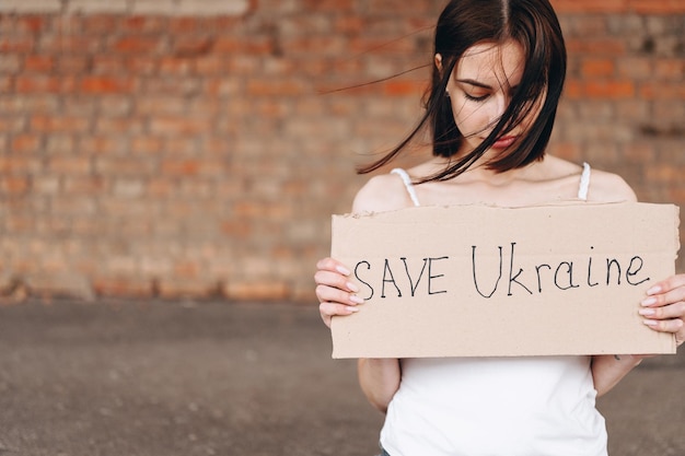 Een brunette vrouw met een karton in haar handen vraagt om de zachte baksteenachtergrond van Oekraïne te redden