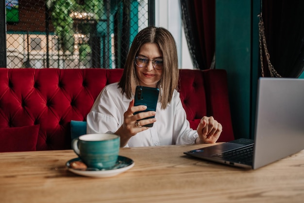 Een brunette zakenvrouw zit aan een tafel in een café met een laptop en kijkt naar haar telefoon