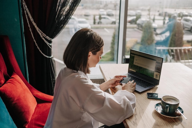 Een brunette zakenvrouw zit aan een tafel in een café met een laptop en winkelt online met een plastic kaart