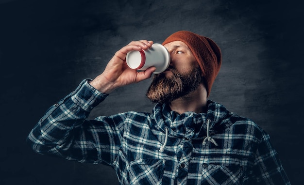 Een brutale bebaarde man gekleed in een hoed en fleece shirt, drinkt koffie uit een papieren glas.
