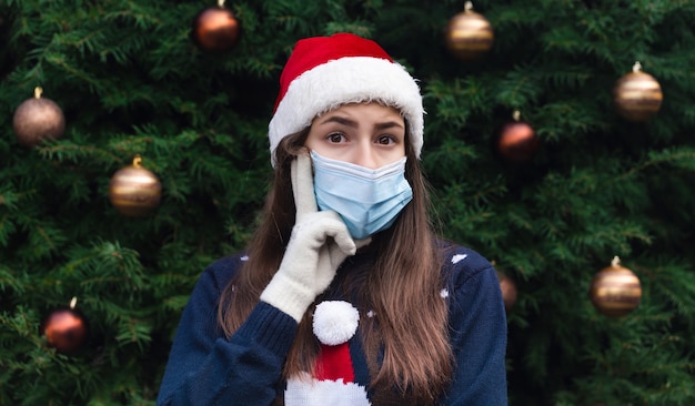 Een cadeau kiezen. Sluit omhoog Portret van vrouw die een hoed van de Kerstman en medisch masker met emotie draagt. Tegen de achtergrond van een kerstboom. Coronapandemie