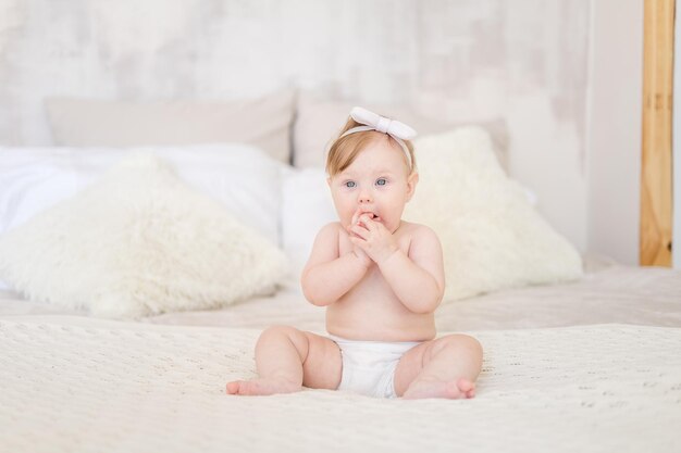 Een charmant babymeisje in luiers zit in een lichte slaapkamer op een bed thuis Slaapkamertextiel