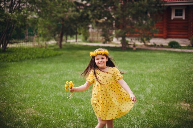 Een charmant meisje in een gele jurk danst met een krans van gele bloemen op haar hoofd op een grasveld van gr...