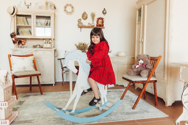 Een charmant meisje in een jurk van natuurlijk materiaal rijdt op een hobbelpaard in het antieke kinderkamerinterieur van haar kamer