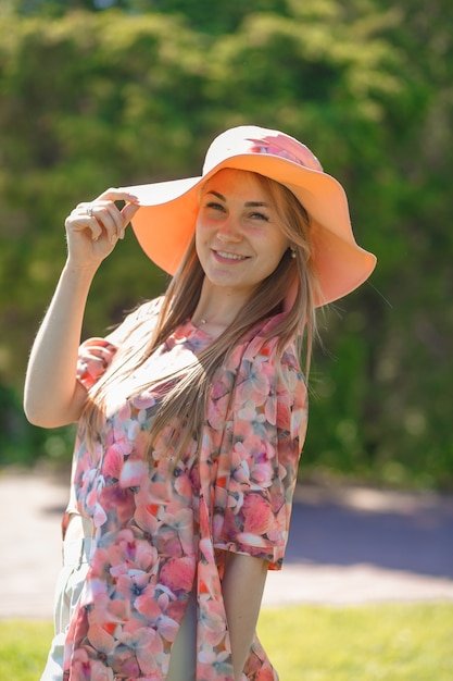 Een charmant meisje in een lichte zomerjurk en een pareohoed loopt in een groen park.
