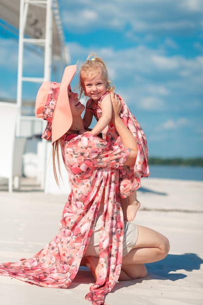 Foto een charmant meisje in een lichte zomerjurk loopt met haar dochtertje over het zandstrand. geniet van warme zonnige zomerdagen.