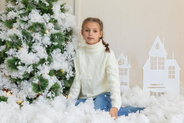 Een charmant meisje in een witte gebreide trui zit tegen de achtergrond van een nieuwjaarsinterieur