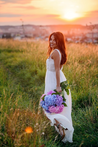 Een charmant meisje in een witte jurk met een boeket tedere gekleurde bloemen loopt in het gazon bij zonsondergang.