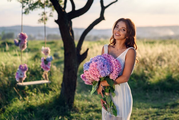 Een charmant meisje met een mooie glimlach in een witte jurk met een boeket tedere gekleurde bloemen bij zonsondergang.