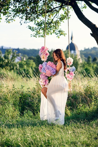 Een charmant meisje met een mooie glimlach in een witte jurk zittend op een houten schommel met bloemen bij zonsondergang.