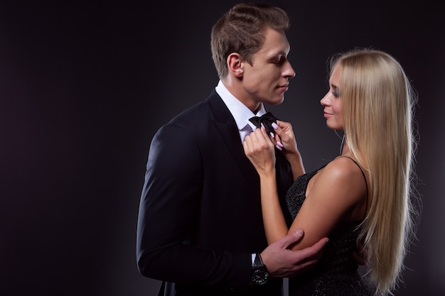 Een charmante blonde vrouw in een zwarte avondjurk knoopt liefdevol een stropdas voor haar man