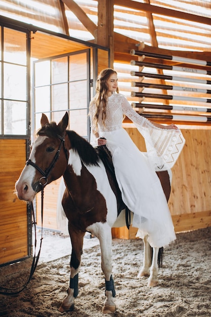 Een charmante boho-bruid berijdt een paard op een ranch bij zonsondergang in de winter