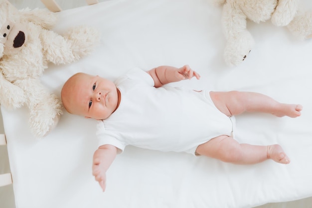 Een charmante glimlachende baby van 2 maanden met blauwe ogen in een witte bodysuit ligt in een wieg naast een teddybeer Uitzicht van bovenaf op een pasgeborene
