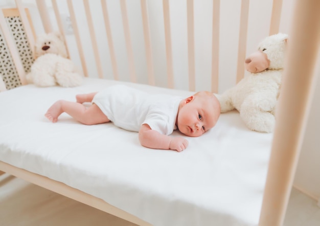 Een charmante glimlachende blauwogige baby van 2 maanden in een witte bodysuit ligt in een wieg naast een teddybeer Uitzicht van bovenaf op een pasgeborene