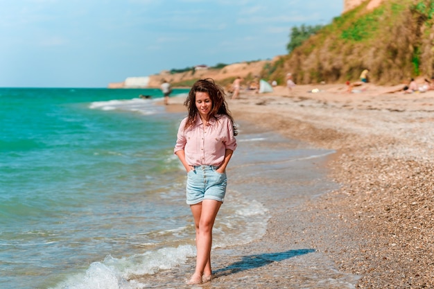 Een charmante jonge vrouw met lang haar en gekleed in een shirt loopt op een zonnige dag langs het strand