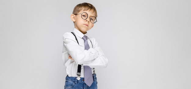 Een charmante jongen in een wit overhemd, bretels, een stropdas en lichte jeans staat. De jongen sloeg zijn armen over zijn borst