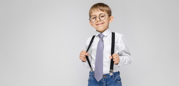 Een charmante jongen in een wit shirt, bretels, een stropdas en lichte jeans staat. de jongen in glazen trok aan de bretels