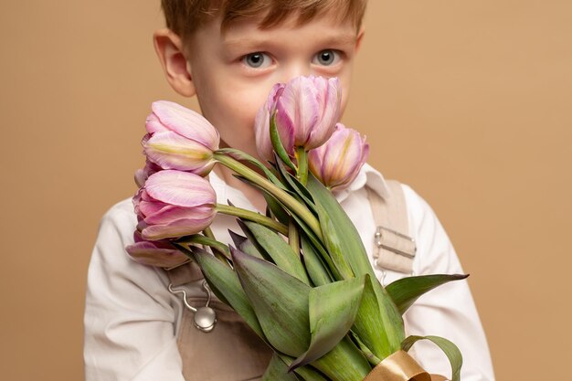 een charmante vierjarige jongen in een beige overall en een wit overhemd houdt een boeket roze tulpen vast