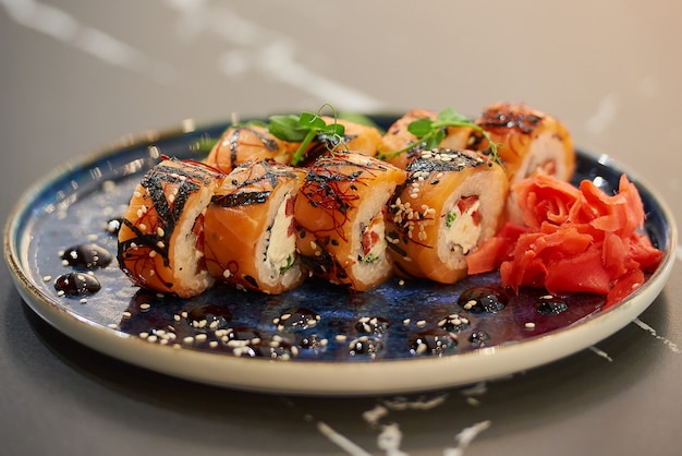Een close-up foto van verse sushi rolt met zalm, Philadelphia kaas en tonijn op een blauwe keramische plaat met saus, sesam, lobules van kumquat, wasabi en bladeren. Zwarte stenen tafel.