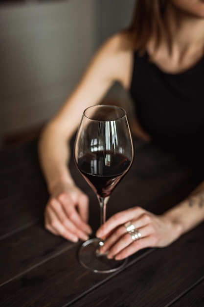 Een close-up van een glas rode wijn in de handen van een blanke jonge vrouw die aan de houten tafel zit