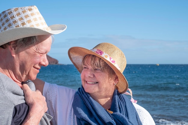 Een close-up van twee lachende senioren die elkaar teder aankijken tijdens de wintervakantie aan zee. Staande op het strand op een zonnige en winderige dag met horizon over het water en de berg