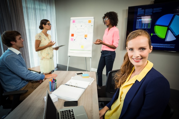 Een collega die bij camera glimlacht terwijl collega's stroomschema op whiteboard bespreken