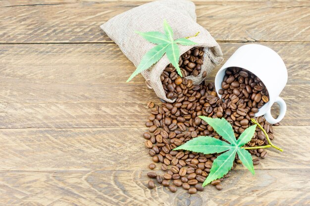 Een compositie van een koffiekopje gevuld met koffiebonen en een zak koffiebonen op een bruine houten ondergrond met groene morijuana-bladeren. bovenaanzicht.